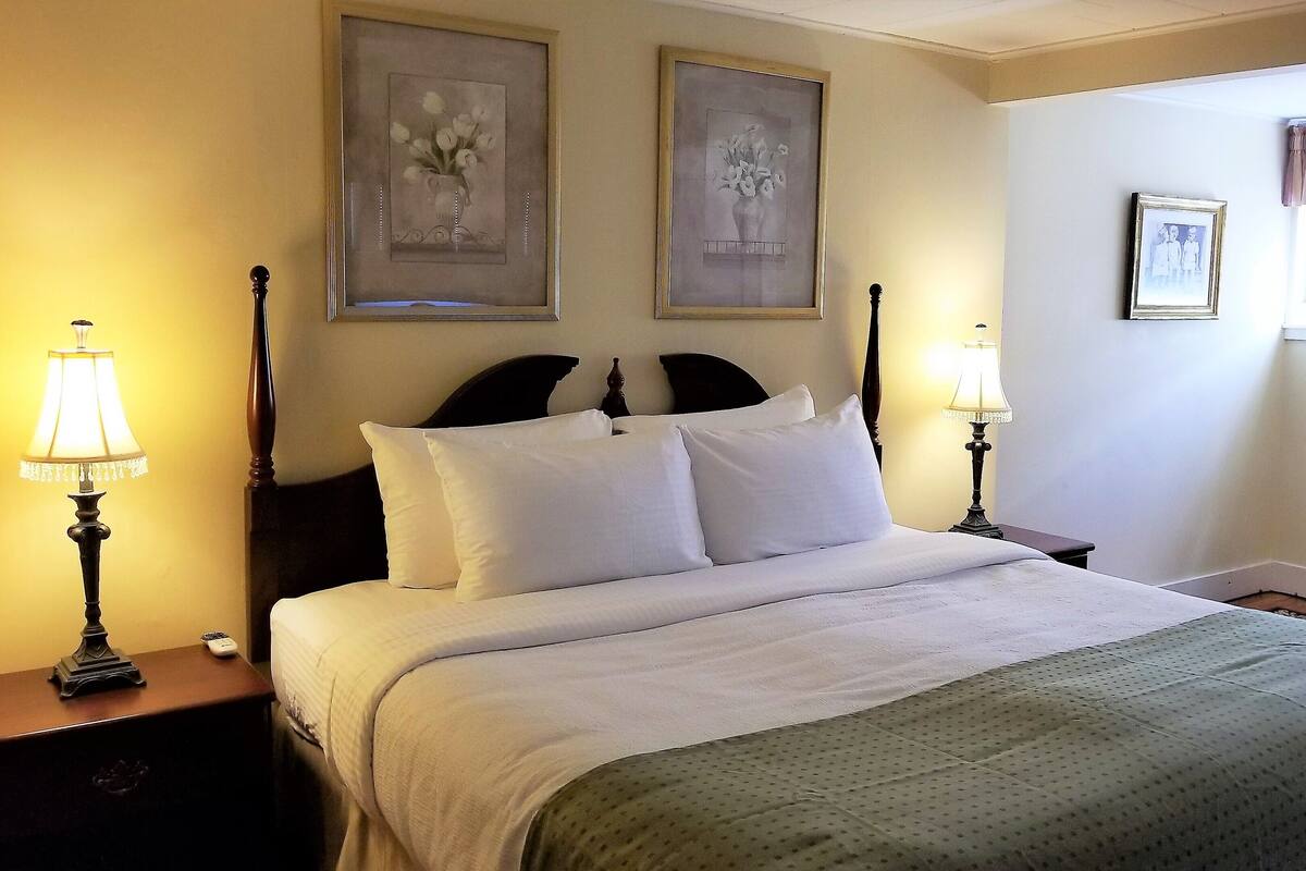 Una cama bien tendida con sábanas blancas, dos lámparas fijadas a la pared y dos cuadros enmarcados con motivos florales encima.