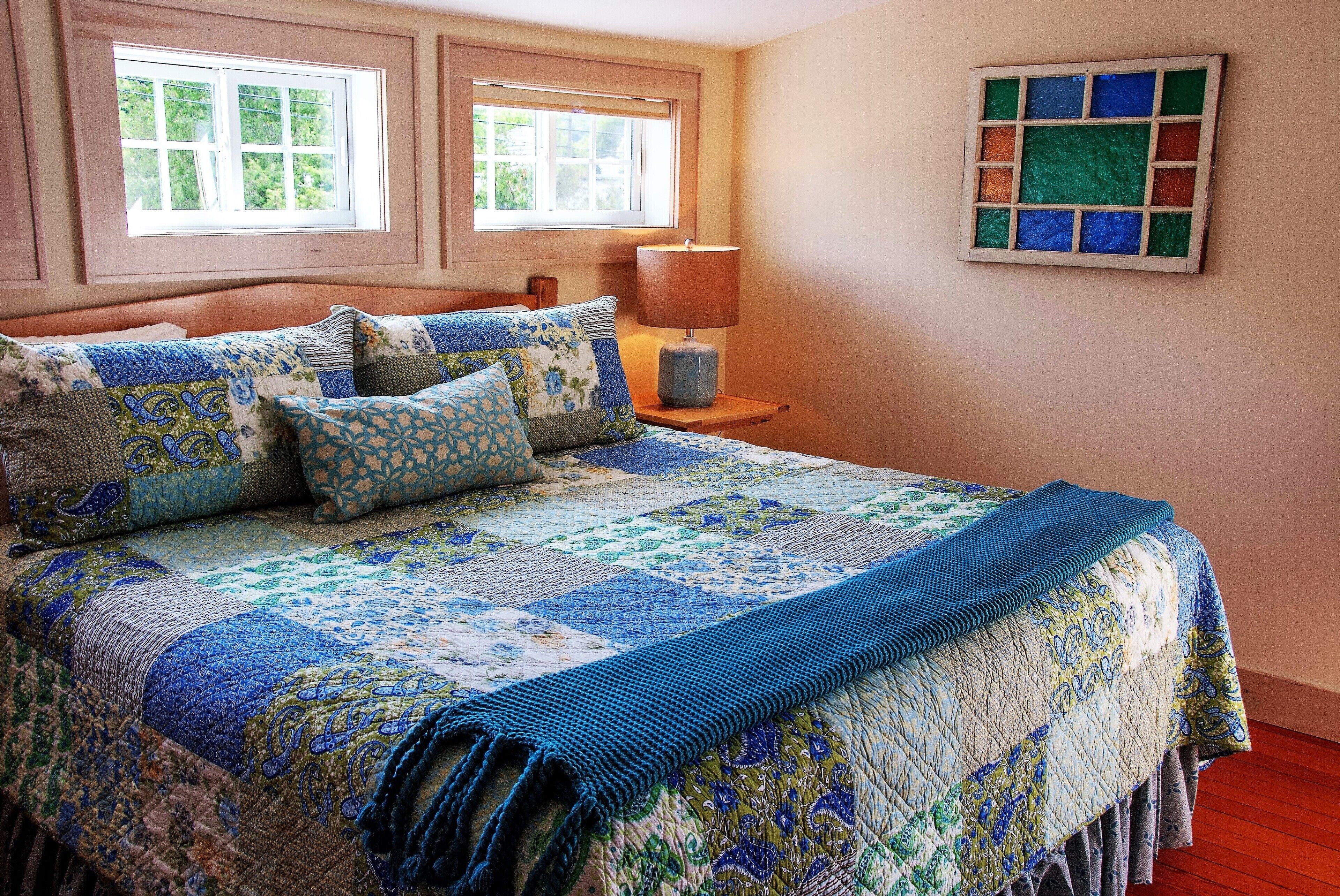 A bed with a blue and green quilt, a bedside lamp, and a window with a view of greenery.