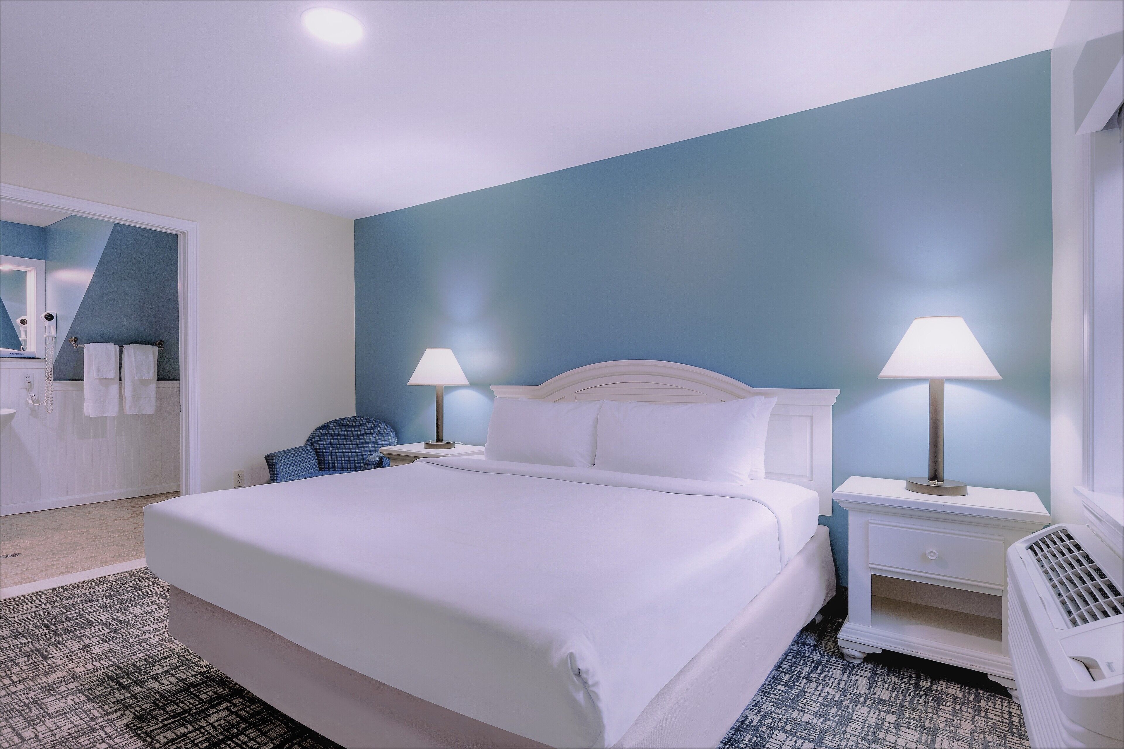 A hotel room with a large bed, two bedside lamps, a white nightstand, a blue chair, and a bathroom visible through an open door.