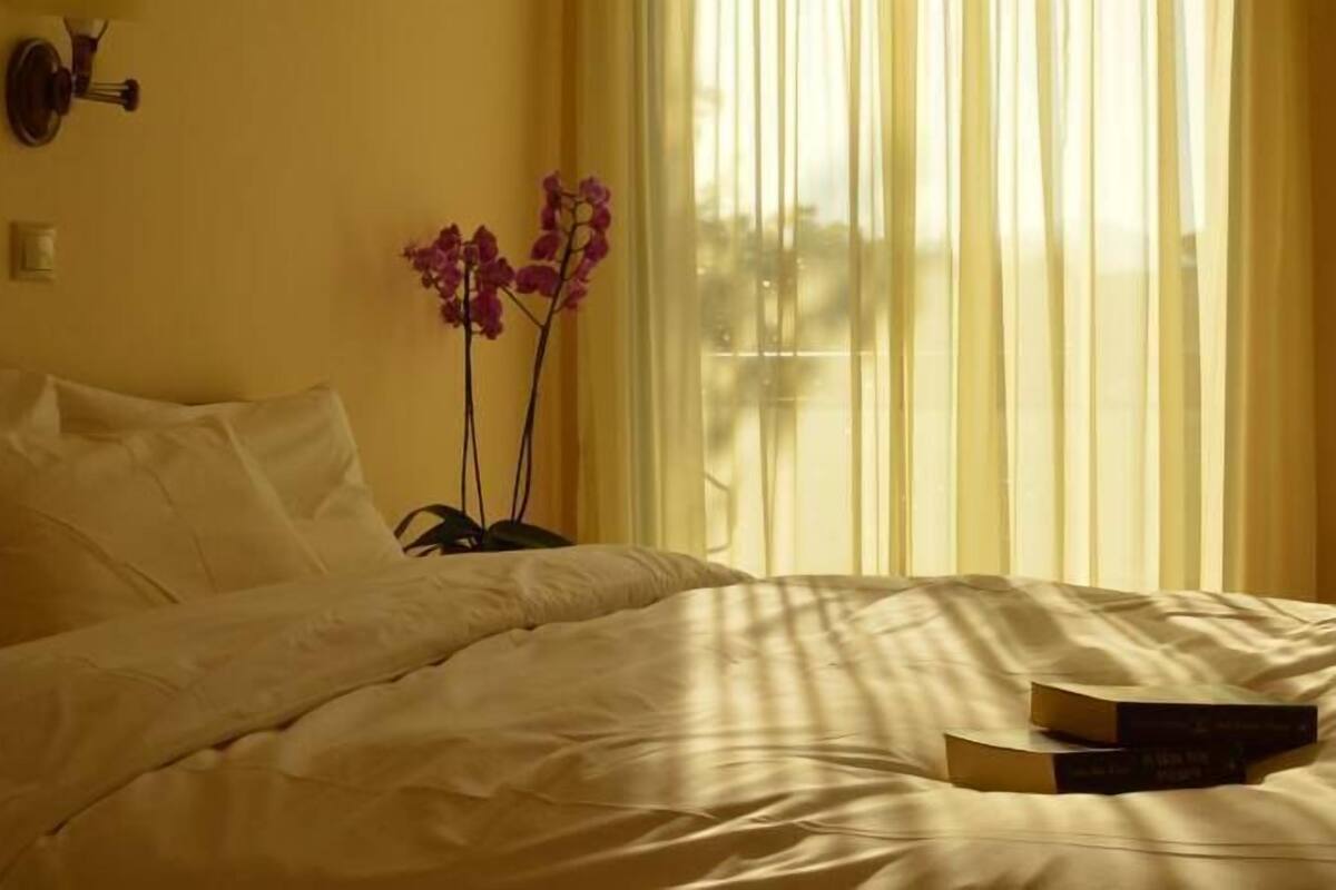 A bed with white bedding, a vase with purple flowers, and two books on the bed.