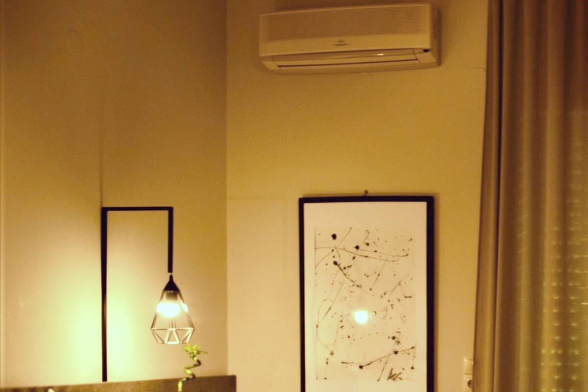 A hotel room with a bed, a nightstand, a lamp, an air conditioning unit, and a framed abstract artwork on the wall.