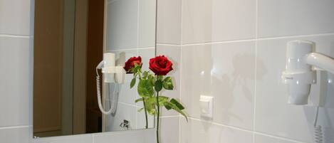 A bathroom with a white sink, a mirror, a hairdryer, and a vase with a red rose.