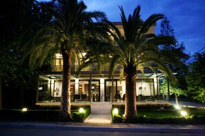 A modern hotel with a glass facade, palm trees, and a well-lit entrance.