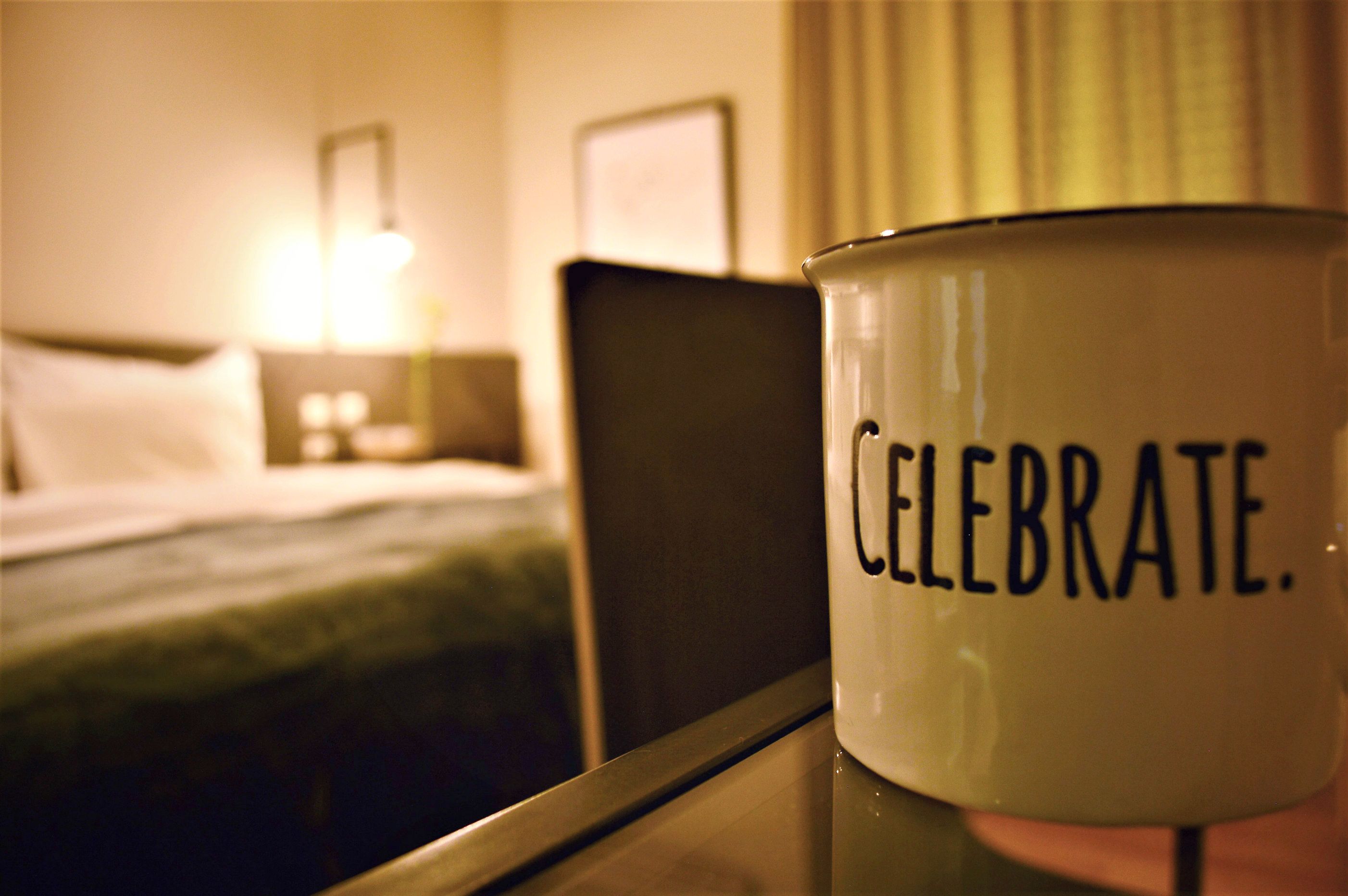 A hotel room with a bed, a nightstand, and a bedside table with a mug that says "CELEBRATE."