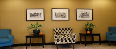 Un espace d’accueil avec trois photographies en noir et blanc encadrées accrochées au mur, un fauteuil à motifs et deux fauteuils bleus, chacun accompagné d’une table d’appoint sur laquelle sont posées des plantes en pot.