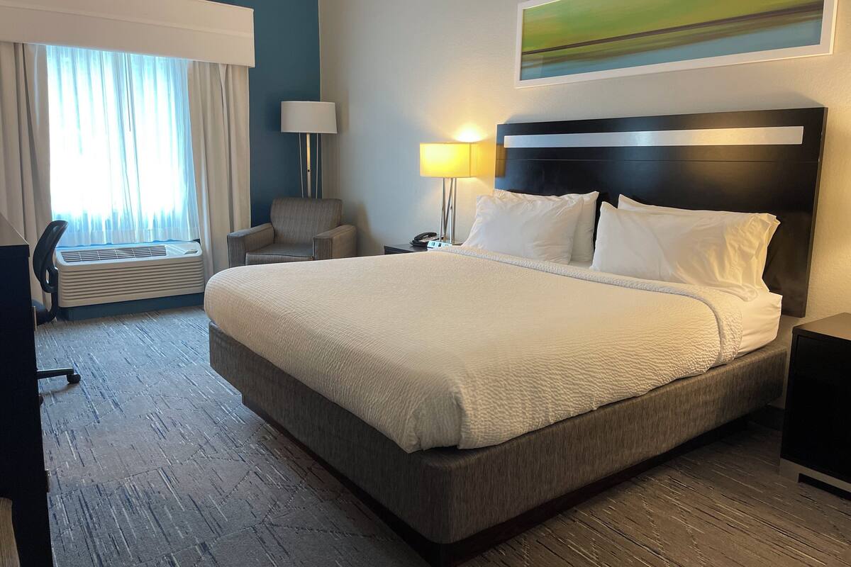 A hotel room with a large bed, a chair, a desk, a lamp, and a painting on the wall.