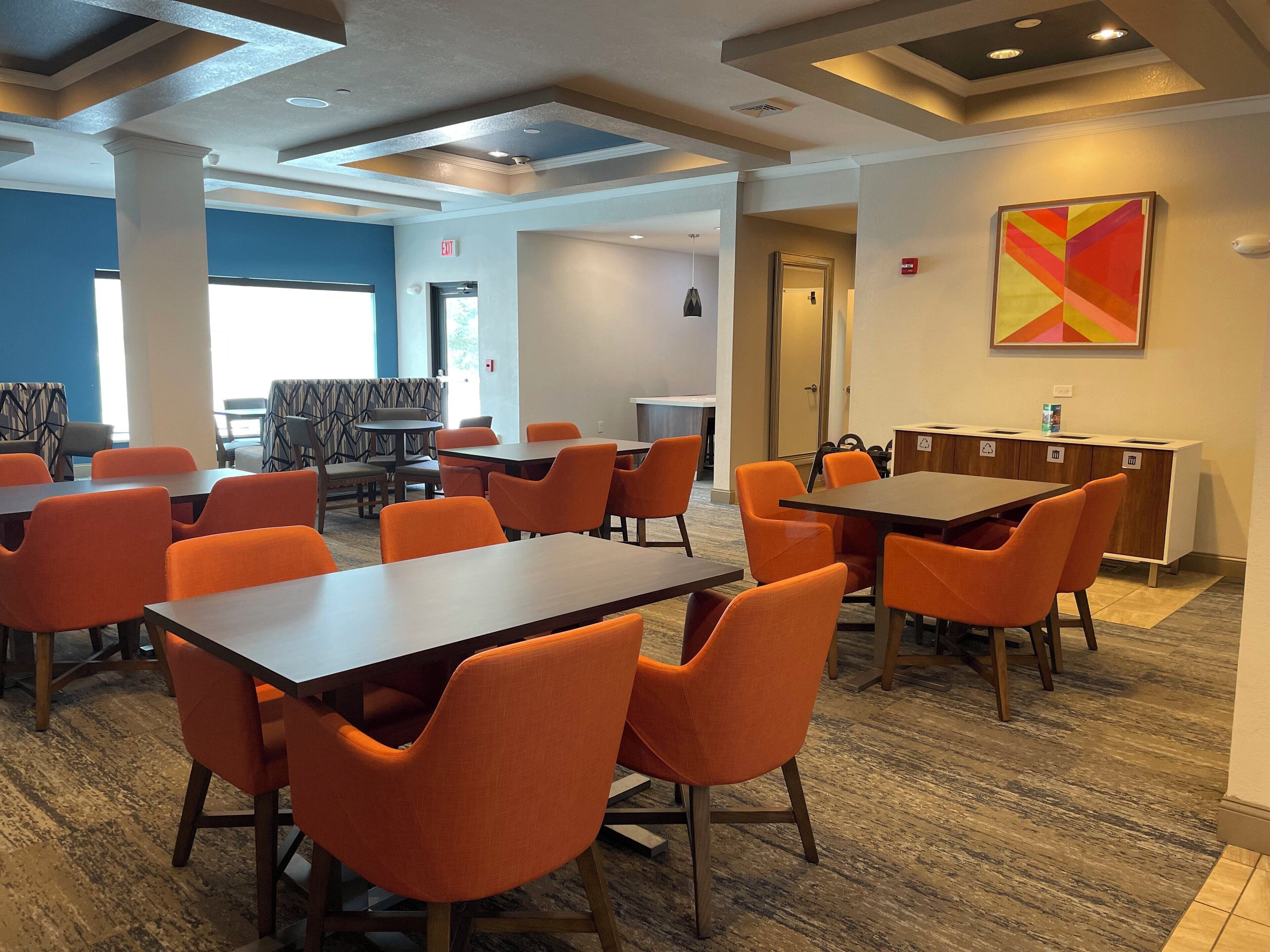 A modern dining area with orange chairs and dark tables, featuring a blue accent wall and a colorful abstract painting.