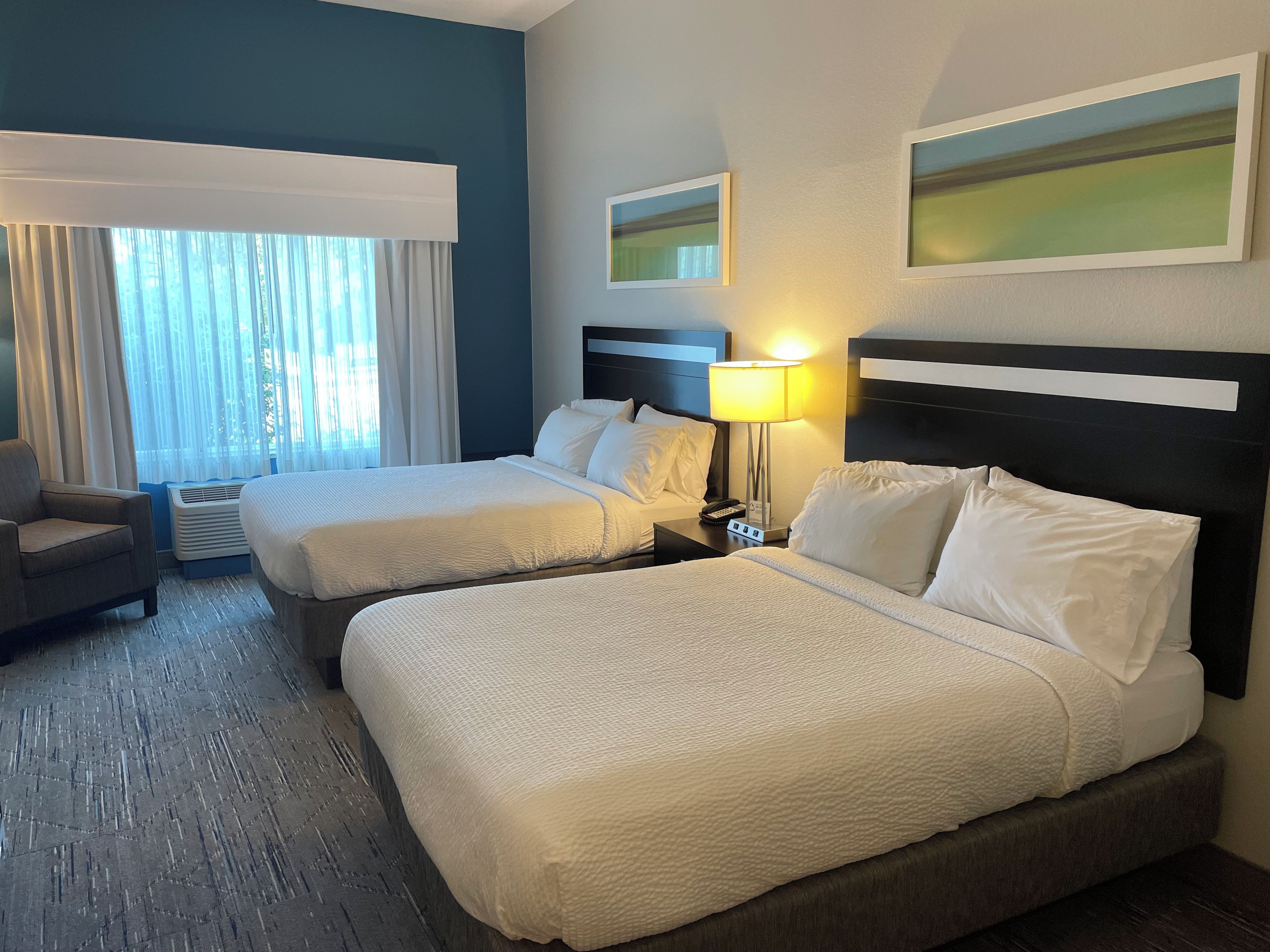 A hotel room with two beds, a chair, a lamp, and a window with curtains.