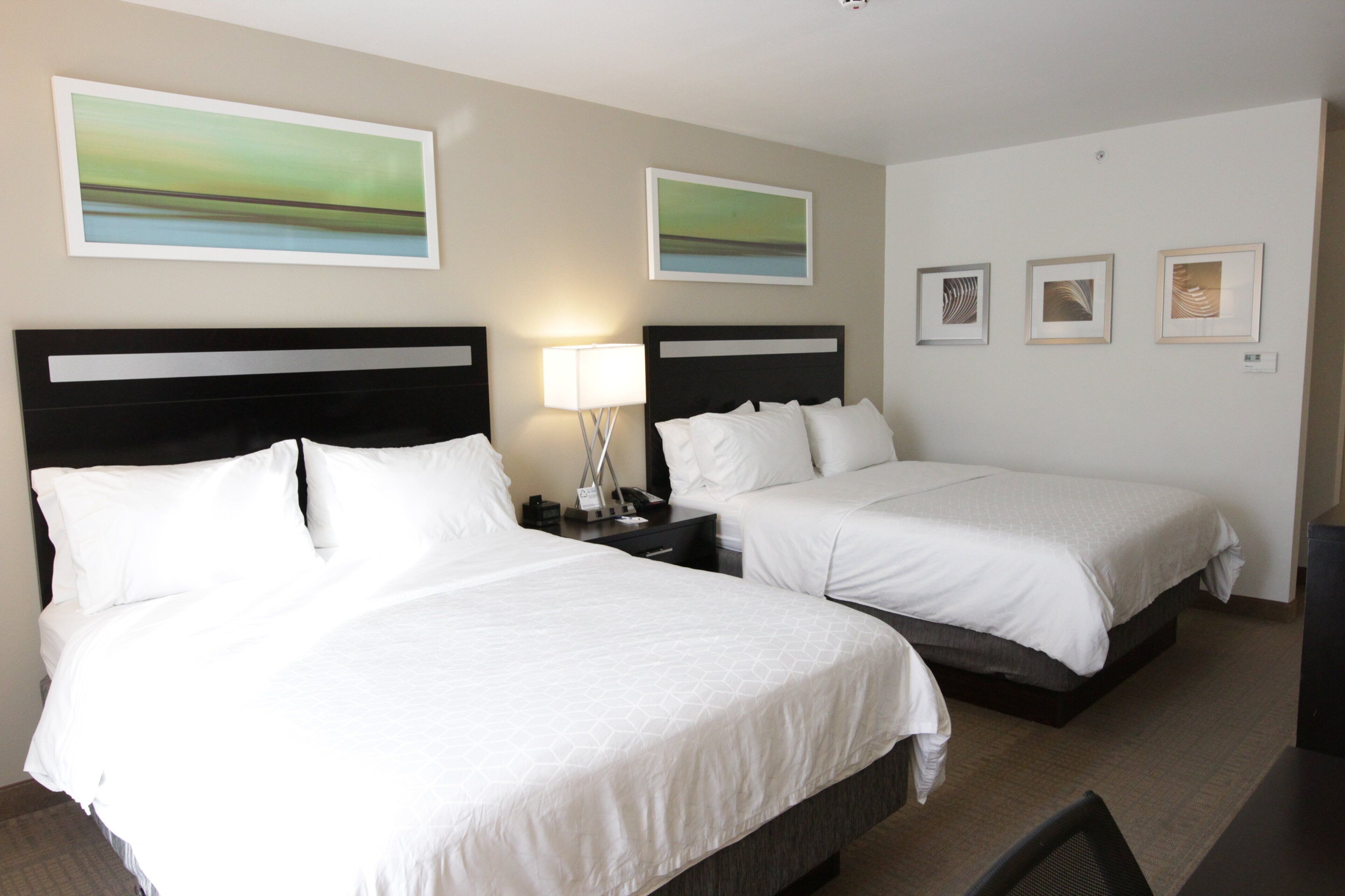 Habitación de hotel con dos camas, una mesita de noche con lámpara y tres cuadros enmarcados en la pared.