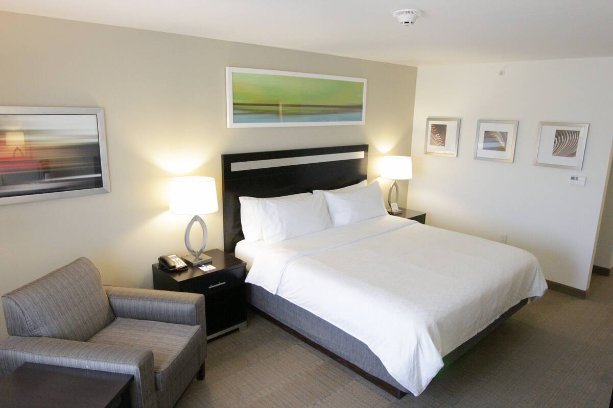 A hotel room with a large bed, a grey armchair, a nightstand with a lamp, and a painting on the wall.