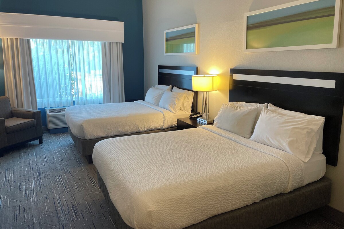 Een hotelkamer met twee bedden, een stoel, een lamp en een raam met gordijnen.