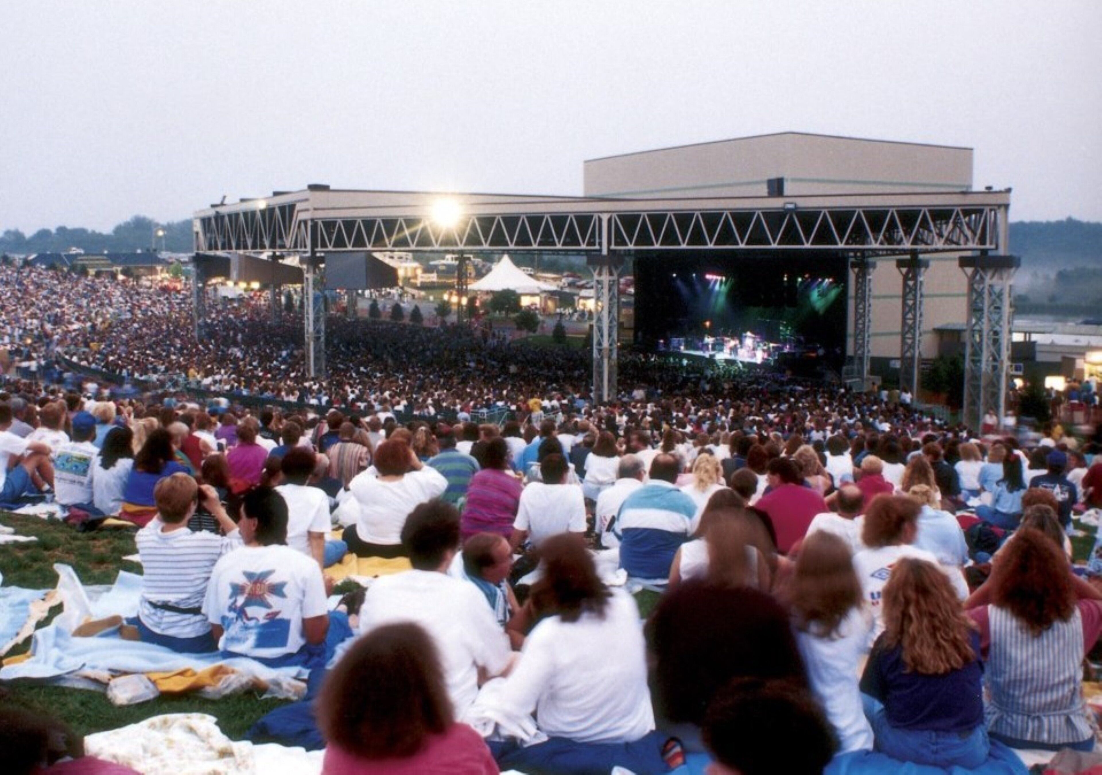 Un amplio recinto para conciertos al aire libre con escenario y público sentado sobre el césped.