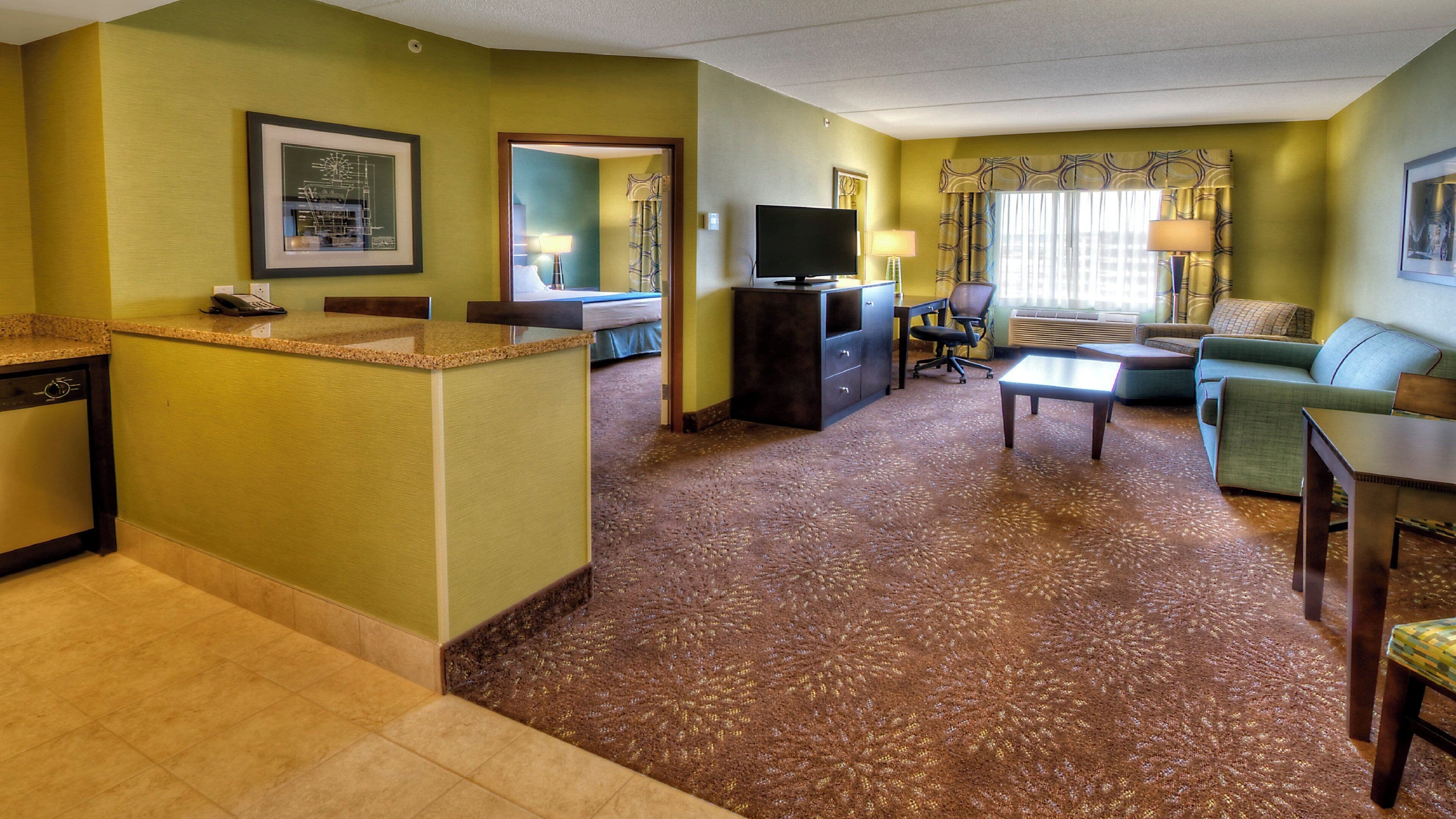 A hotel room with a kitchenette, a living area with a sofa and a coffee table, a television, and a large window with curtains.