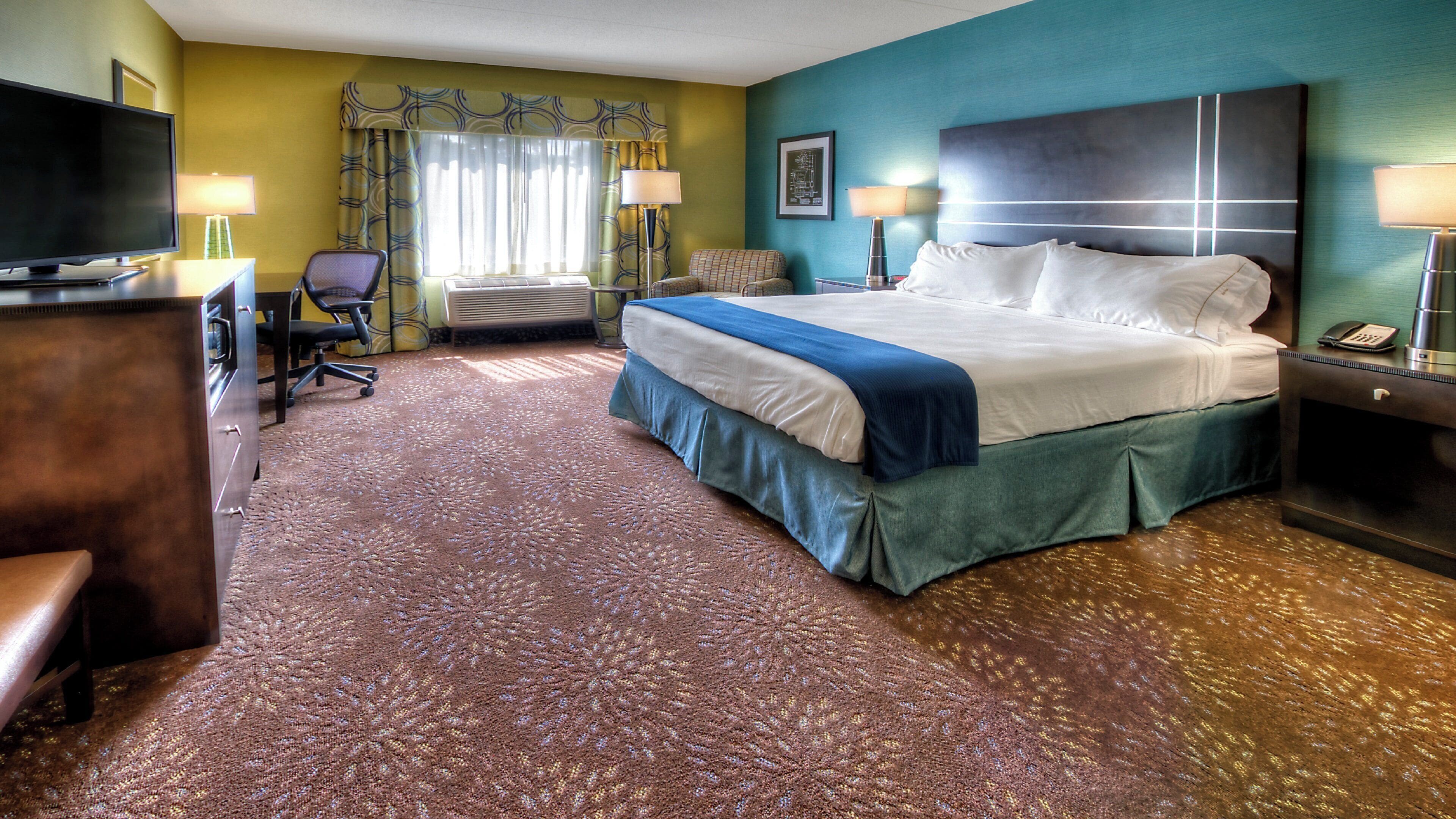 A hotel room with a bed, a desk, a chair, a television, and a nightstand with a lamp.