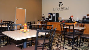 A hotel dining area with tables and chairs, a kitchen in the background, and a wall with the hotel's name.