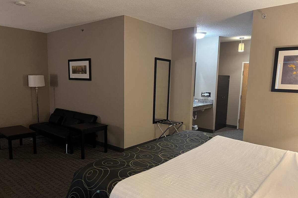 Habitación de hotel con cama, escritorio, silla, lámpara, espejo y baño visible a través de una puerta abierta.
