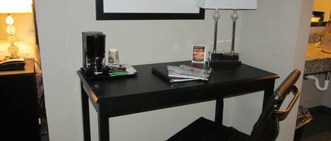 A hotel room with a small desk, a chair, a lamp, a coffee maker, and a framed picture on the wall.