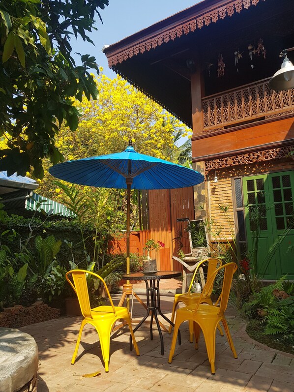 Un patio con sillas amarillas y una sombrilla azul, rodeado de abundante vegetación y un edificio de madera con detalles intrincados.