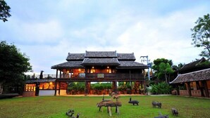 A large wooden building with a balcony, surrounded by a grassy area with animal sculptures. - Lanna Ricebarn (Chiang Mai)