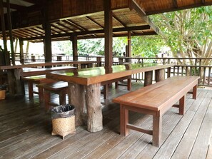 A covered outdoor seating area with wooden tables and benches, surrounded by a wooden fence and trees. - Lanna Ricebarn (Chiang Mai)