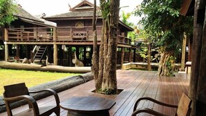 A wooden deck with two chairs and a round table, surrounded by trees and a building with a traditional design. - Lanna Ricebarn (Chiang Mai)