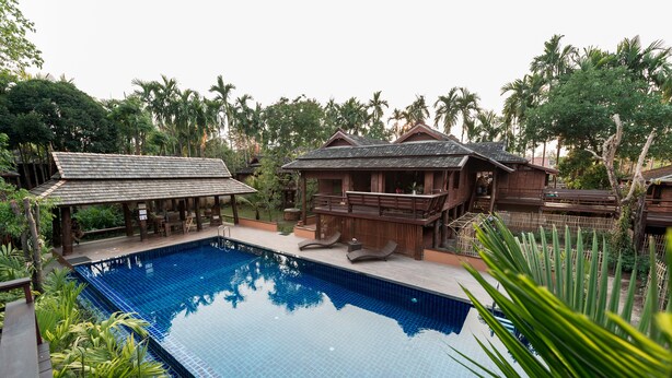 A pool area with wooden structures, including a lounge and a covered seating area, surrounded by lush greenery and palm trees. - Lanna Ricebarn (Chiang Mai)