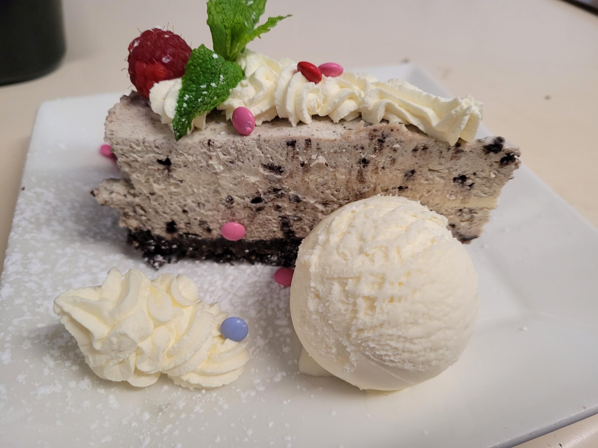 Una porción de pastel de queso con crema batida y una frambuesa encima, acompañada de una bola de helado de vainilla y un toque de crema batida.
