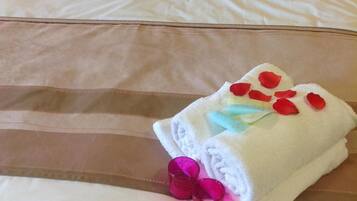Una cama bien hecha con ropa de cama blanca, dos almohadas y una toalla doblada con pétalos de rosa sobre un cabecero de madera.