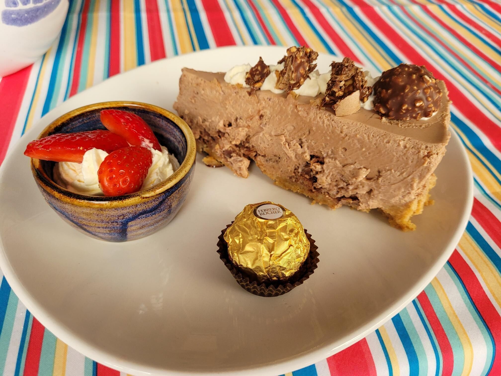A dessert plate with a slice of cheesecake, a small bowl of whipped cream with strawberries, and a Ferrero Rocher chocolate.