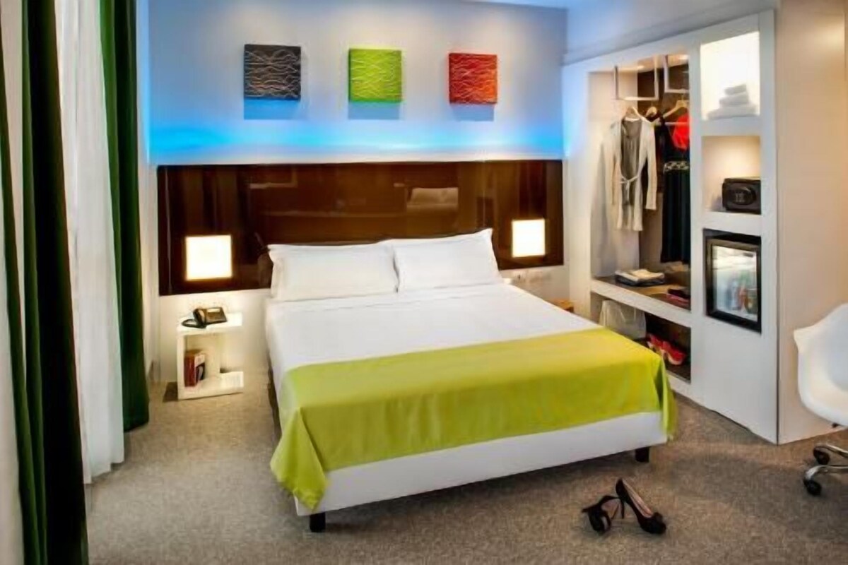 Habitación de hotel con cama, lámparas de noche, escritorio con silla, perchero y televisor.