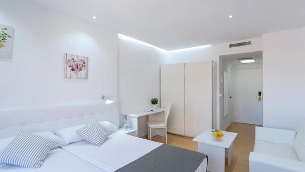 Quarto de hotel moderno com uma cama grande, uma escrivaninha, uma cadeira, uma mesinha com frutas e um sofá branco.