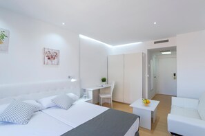 Ein modernes Hotelzimmer mit einem großen Bett, einem Schreibtisch, einem Sessel, einem kleinen Tisch mit Früchten und einem weißen Sofa.