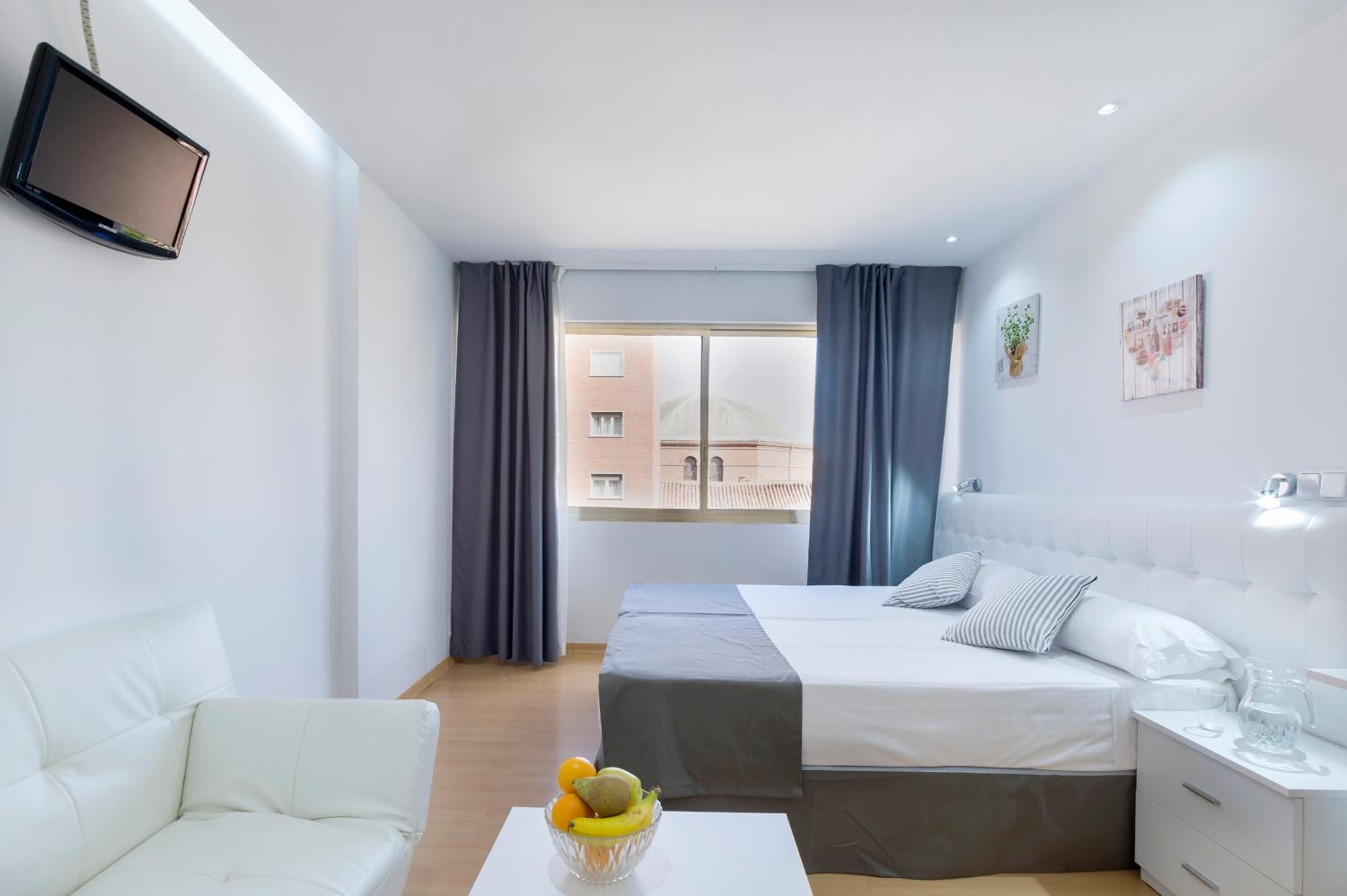 Una habitación de hotel moderna con cama, sofá, televisor y ventana con cortinas.