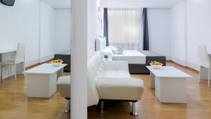 Quarto de hotel moderno com uma cama branca, dois sofás brancos, uma mesa de centro branca e uma pequena mesa lateral com uma tigela de frutas.