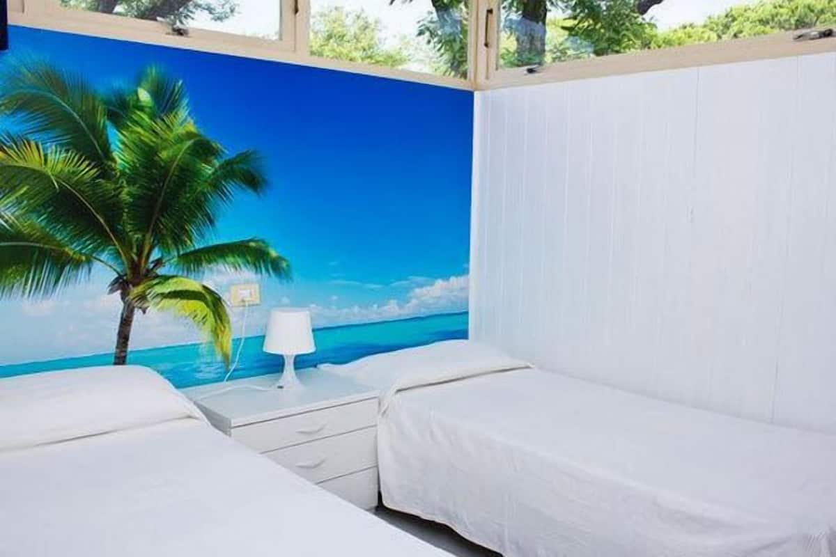 Une chambre avec deux lits, une table de chevet, une lampe et une fresque murale représentant une scène de plage.