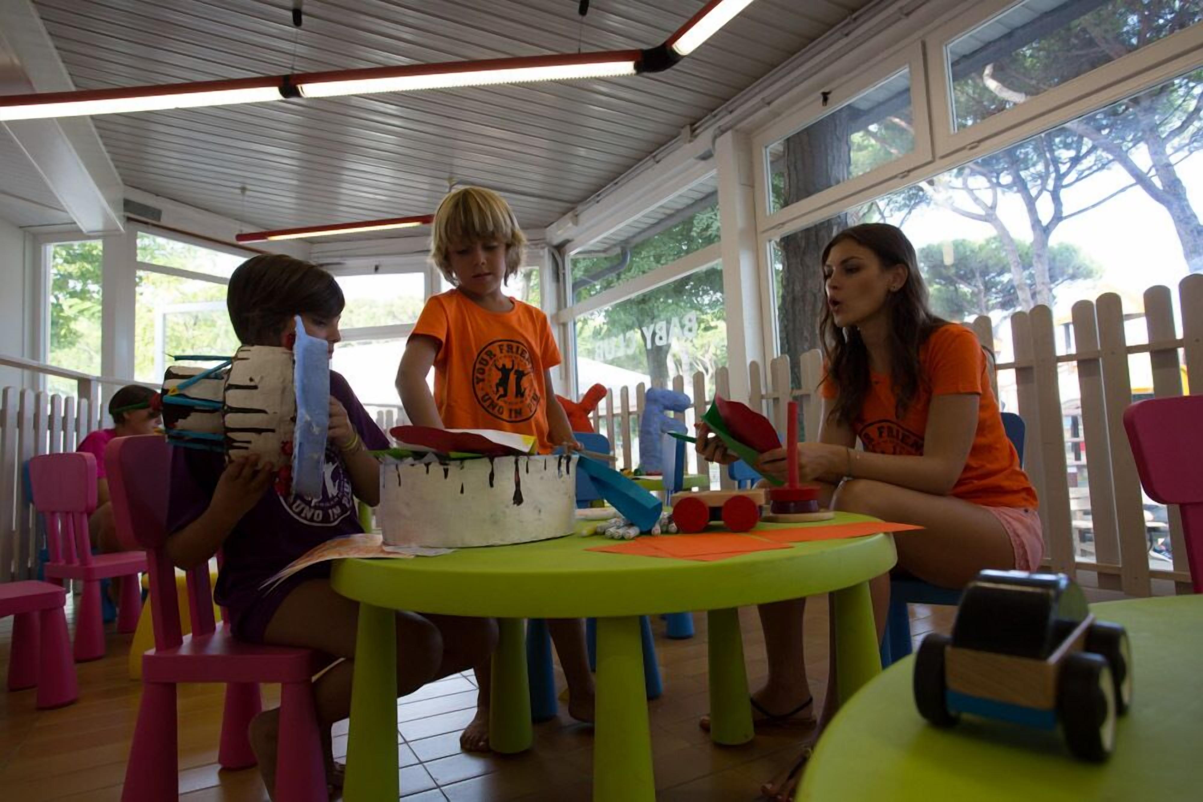 A group of children engaged in a craft activity at a table with a white fence visible through the window.