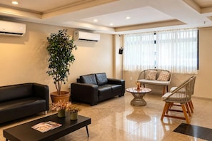 A modern living room with a black leather sofa, a coffee table, a potted plant, and a seating area with a wicker chair and cushions.