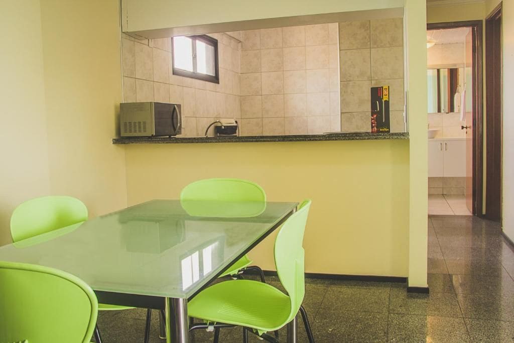 Una habitación con una mesa de vidrio, cuatro sillas verdes, un microondas y una ventana pequeña.