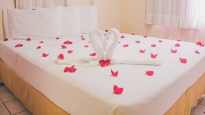 Een net opgemaakt bed met witte lakens en een handdoekje gevouwen in de vorm van een hart, omringd door rozenblaadjes.