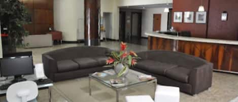 Um lobby moderno com um sofá marrom escuro curvo, uma mesa de centro de vidro com um vaso de flores, um balcão de recepção e uma cadeira de escritório branca.
