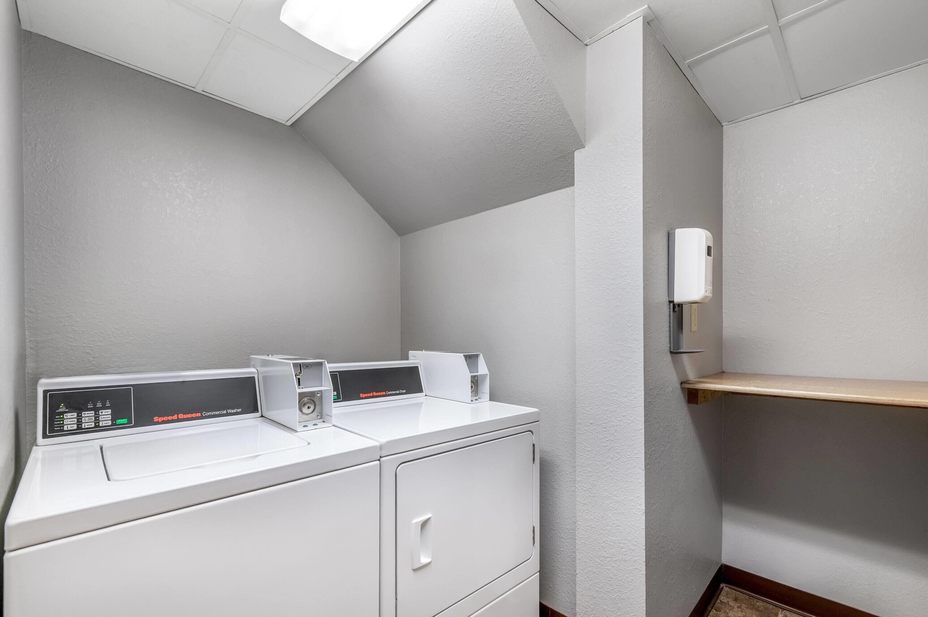 A laundry room with two washing machines and a dryer, a small shelf, and a wall-mounted hand dryer.