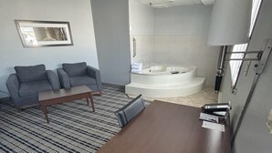 A modern hotel room with a gray sofa, a wooden coffee table, a large mirror, and a corner bathtub. - AmericInn by Wyndham Fergus Falls - Conference Center (Fergus Falls)