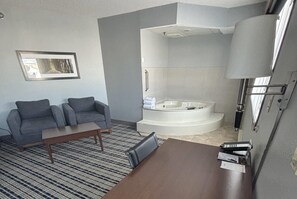 A modern hotel room with a gray sofa, a wooden coffee table, a large mirror, and a corner bathtub.