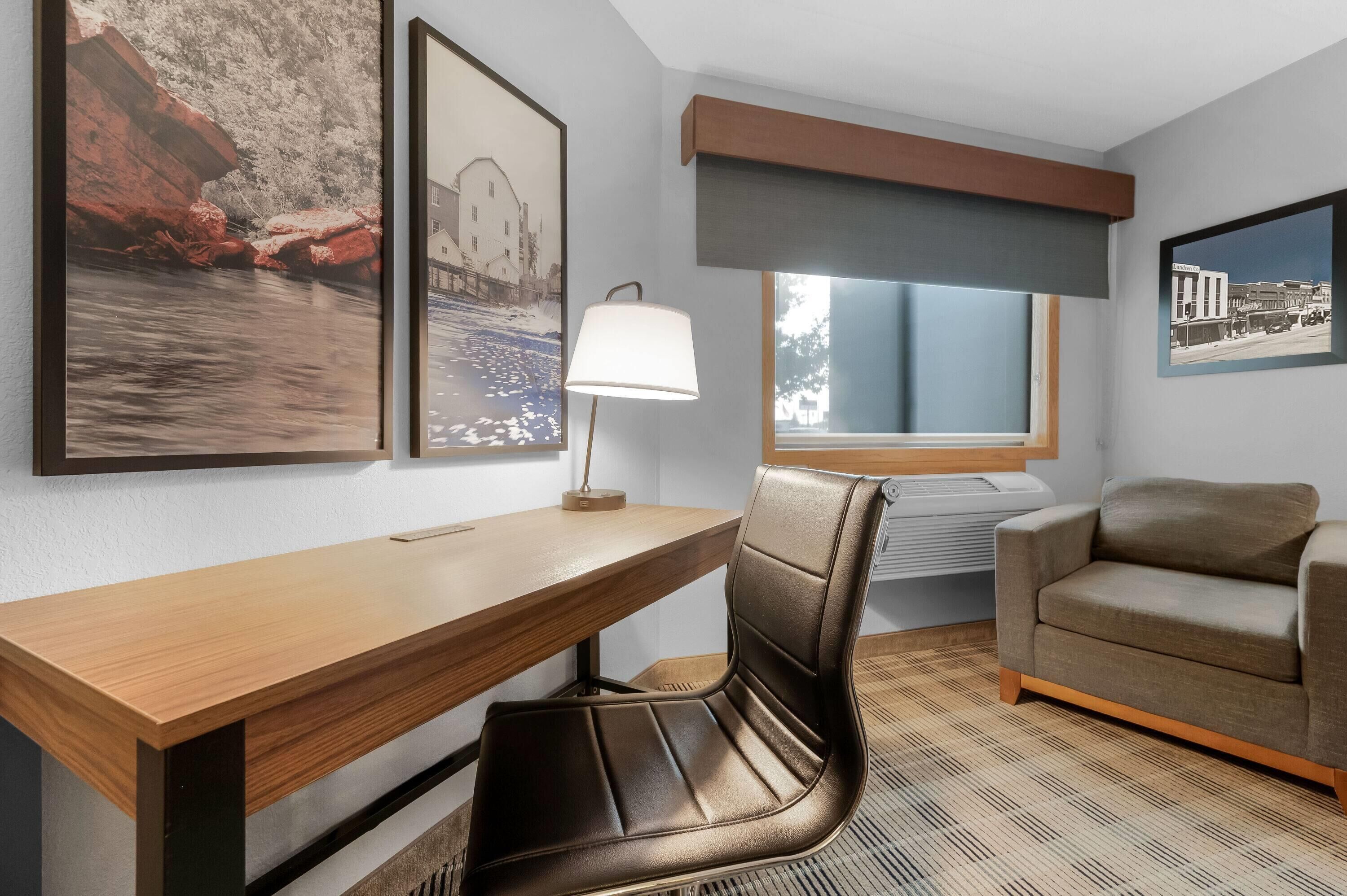 A hotel room with a wooden desk, a leather chair, a lamp, and framed pictures on the wall.