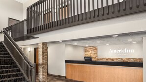 A hotel reception area with a reception desk, a staircase, and a stone pillar. - AmericInn by Wyndham Fergus Falls - Conference Center (Fergus Falls)