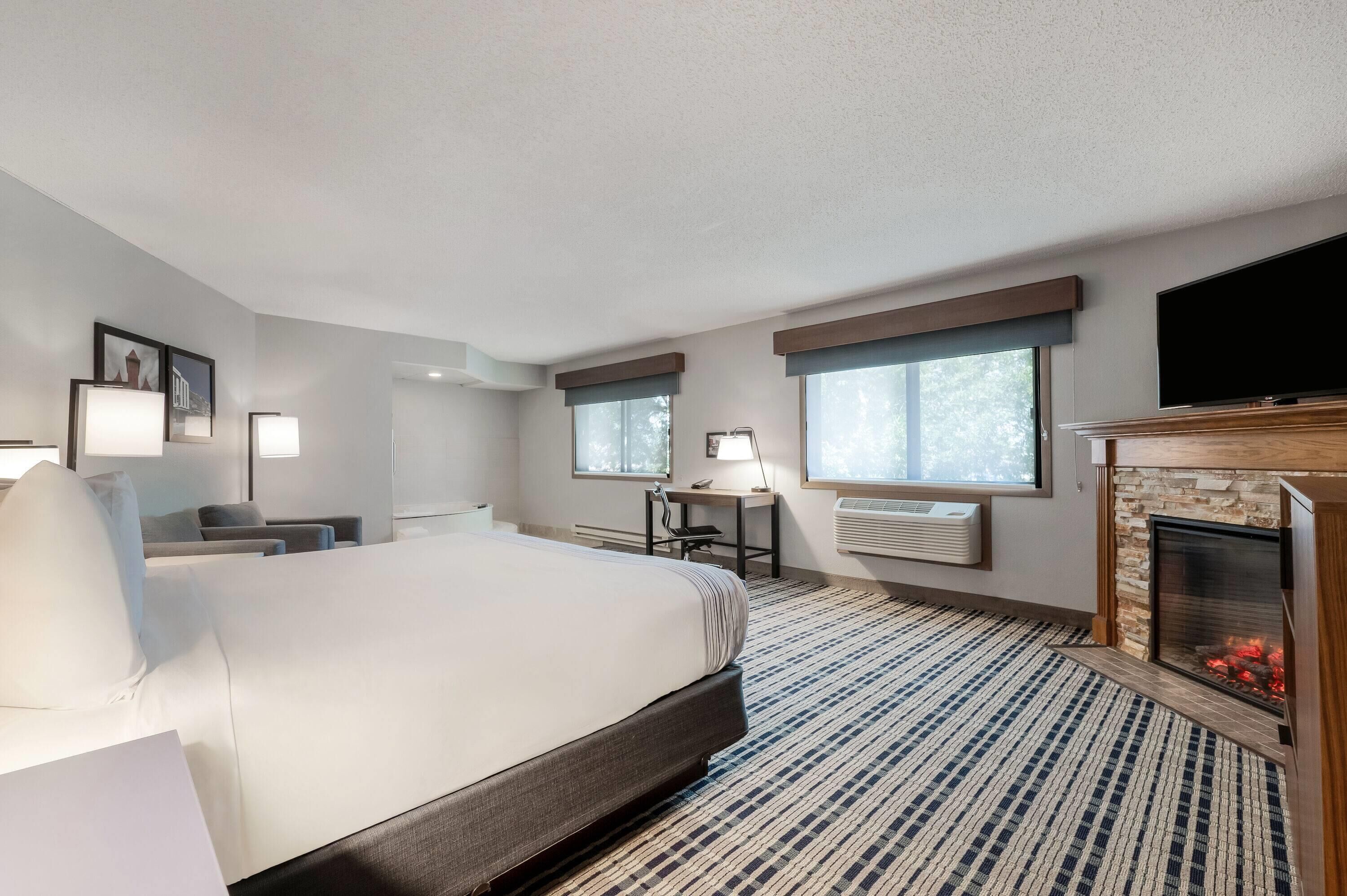 A hotel room with a large bed, a desk, a chair, a flat-screen TV, a fireplace, and two windows.