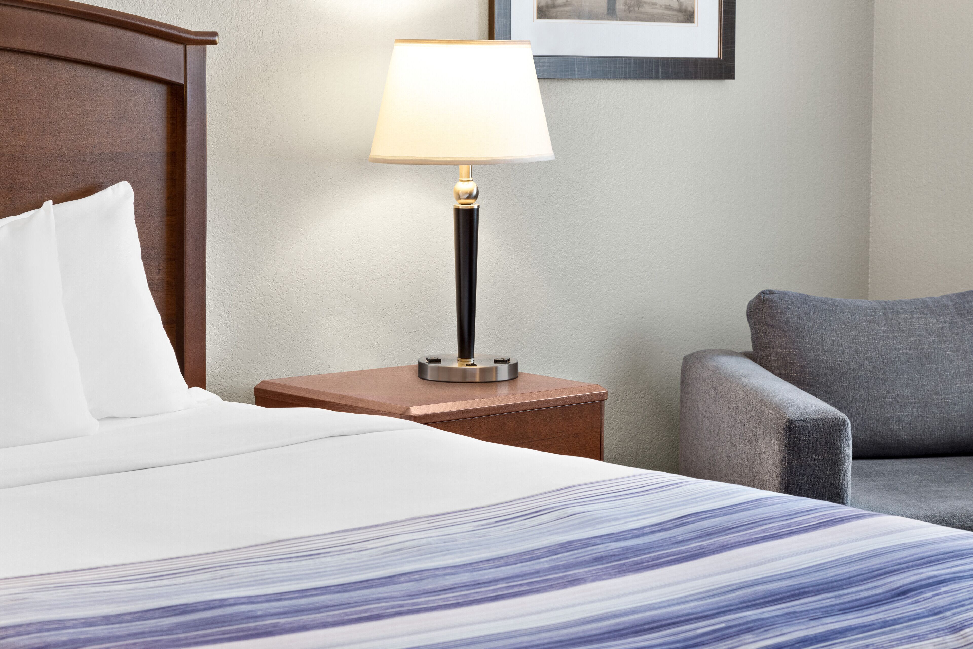 A neatly made bed with a patterned bedspread, a bedside lamp, a gray armchair, and a framed picture on the wall.