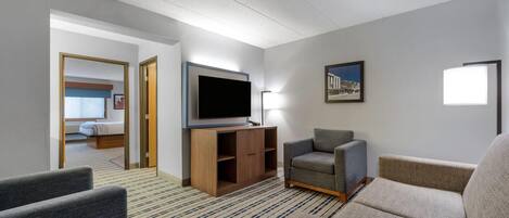 A hotel room with a flat-screen TV, a sofa, a chair, a wooden cabinet, and a patterned carpet.