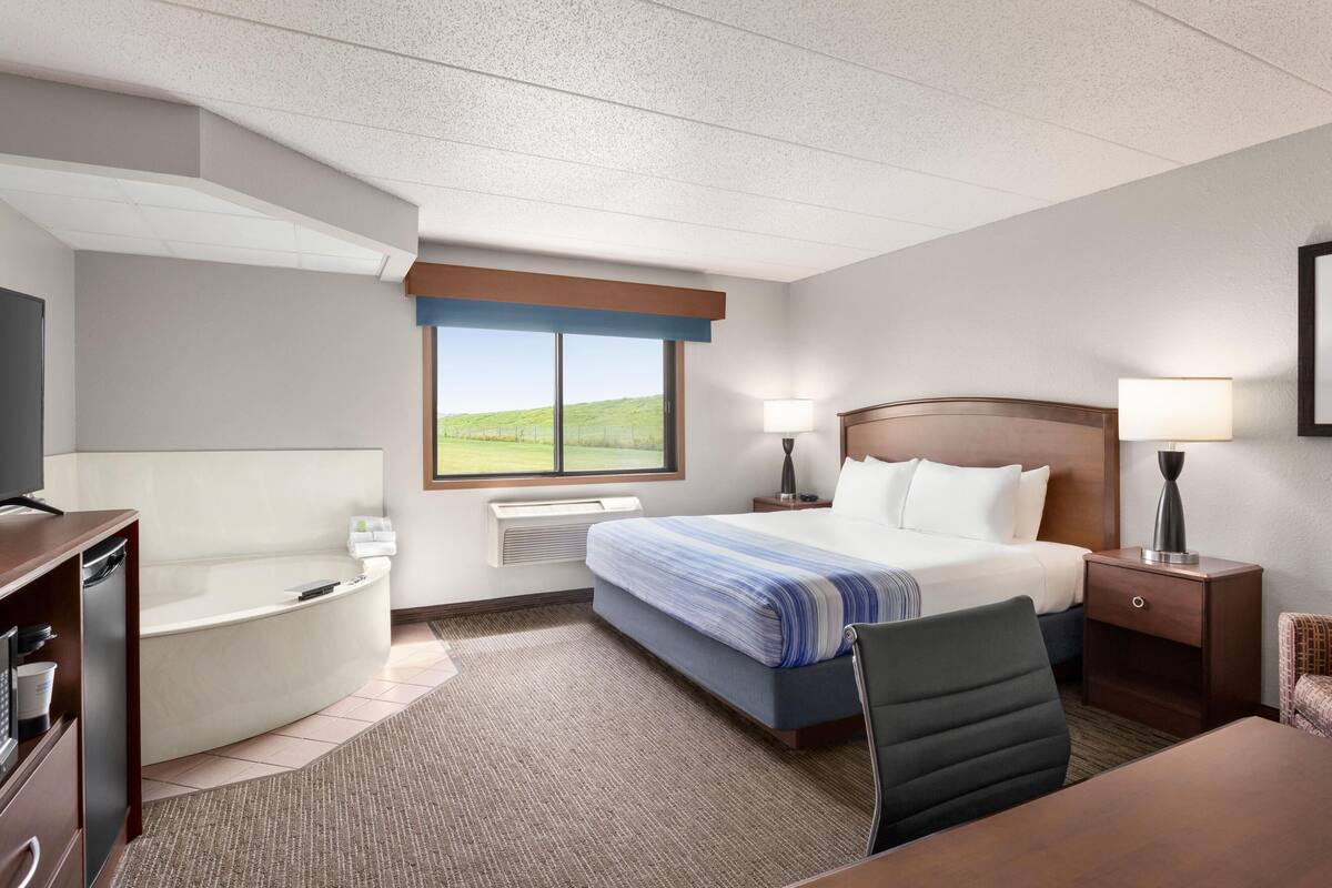 Habitación de hotel con una cama grande, un escritorio, una silla, una lámpara de escritorio, una mesita de noche con lámpara, un televisor, una bañera empotrada y una ventana con vistas a un paisaje verde.
