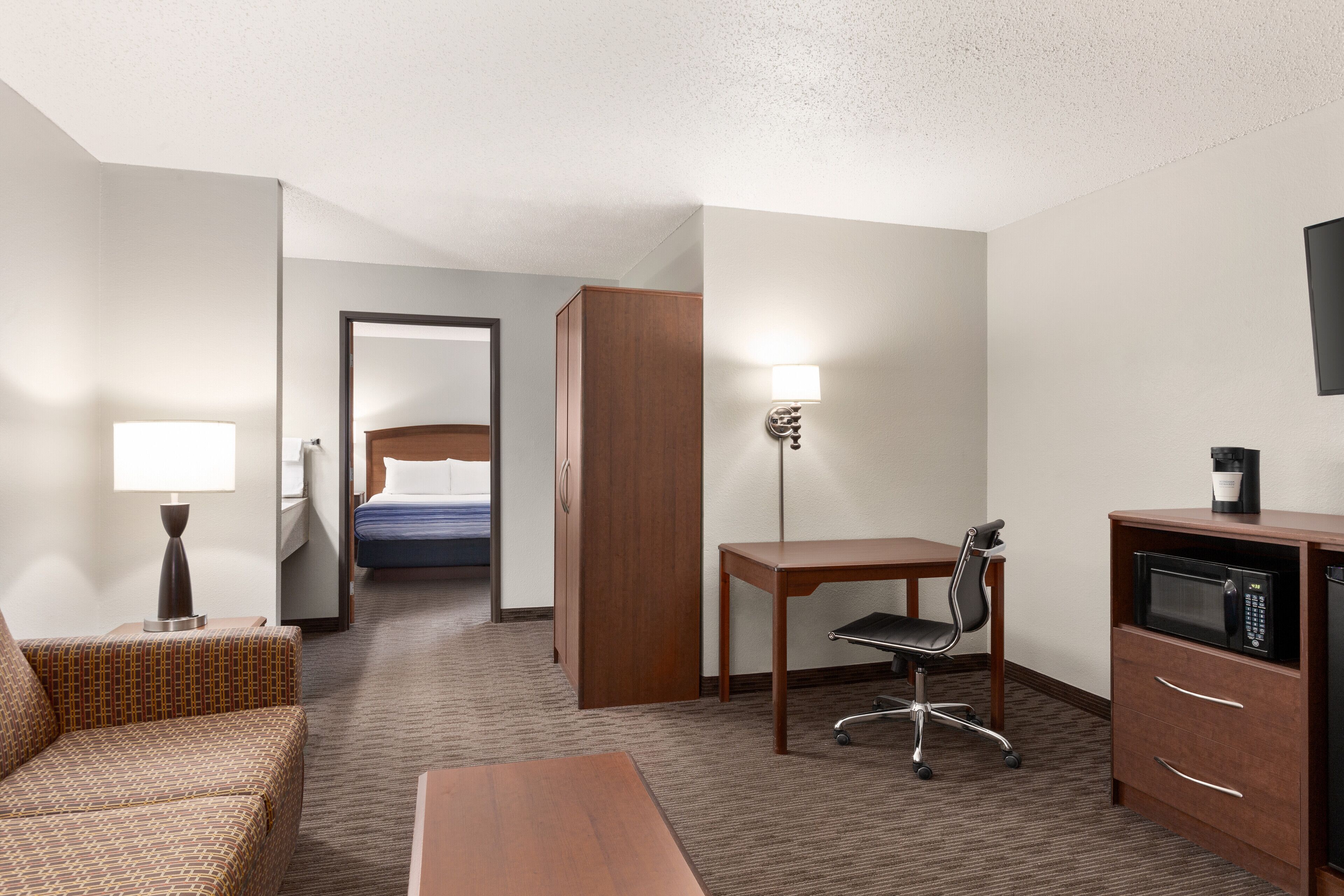 A hotel room with a bed, a desk, a chair, a lamp, a microwave, and a coffee maker.